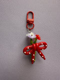 A red plastic keychain bag featuring a large bow with white pearl-like accents, a small white flower, and a red flower charm, attached to a red metal clasp, shown on a gray textured fabric background.