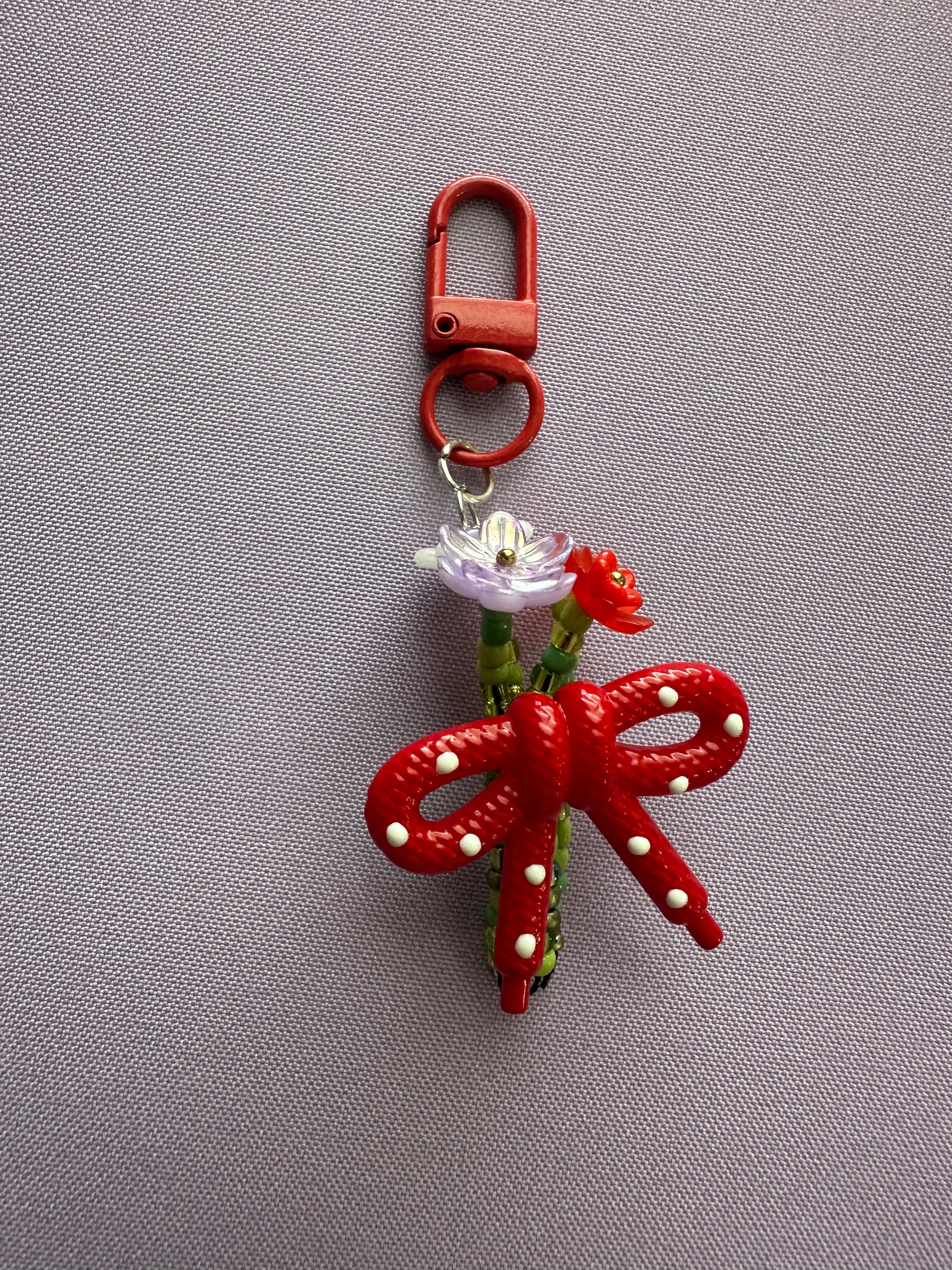 A red plastic keychain bag featuring a large bow with white pearl-like accents, a small white flower, and a red flower charm, attached to a red metal clasp, shown on a gray textured fabric background.