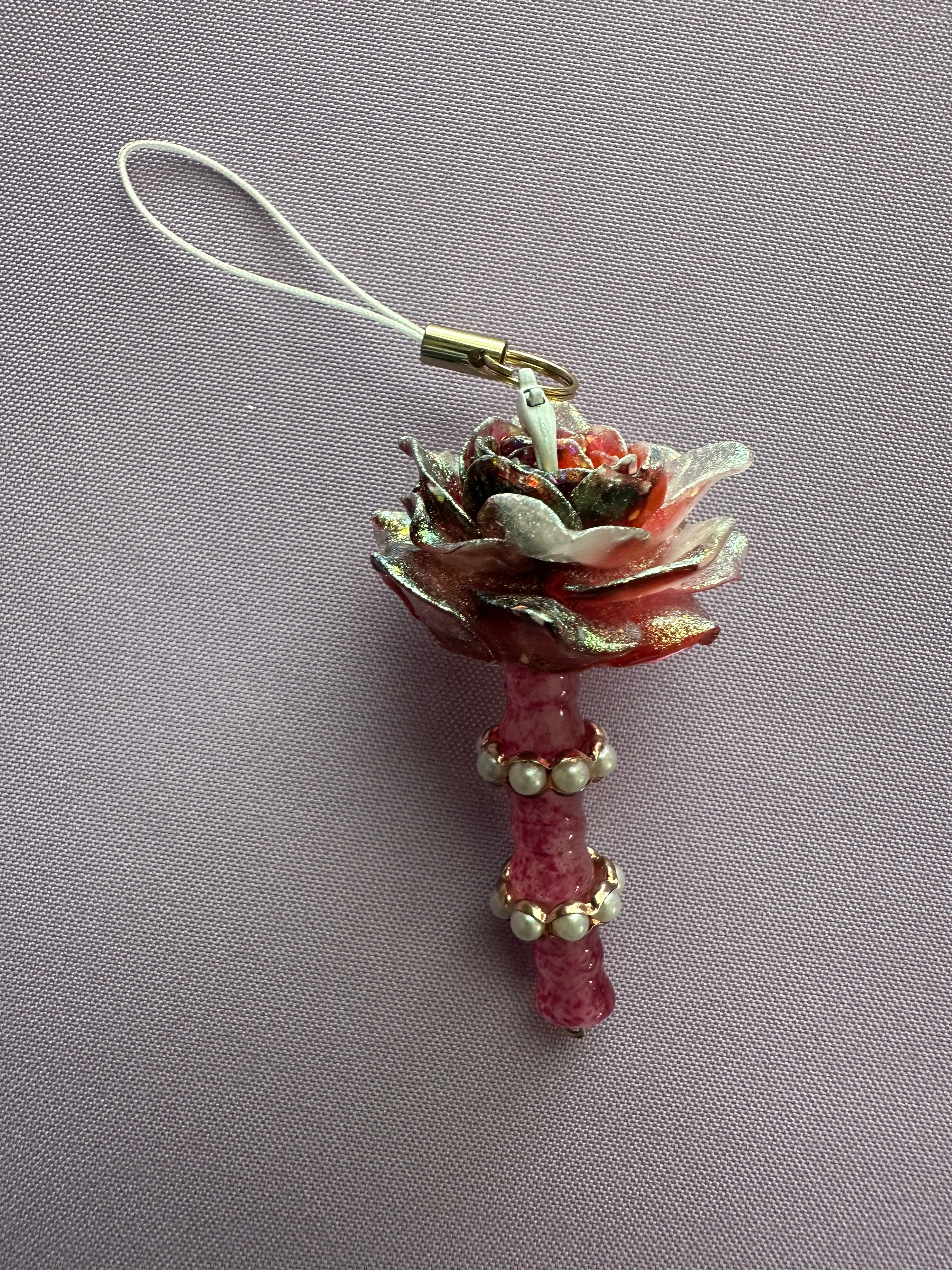 A decorative rose-shaped charm with a white cord loop, featuring layered pink and white petals with metallic gold glitter accents and a pink stem decorated with small white beads, shown on a textured gray background.