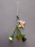 A handmade charm with a white loop strap, a silver clasp, a pink flower, and three dangling beaded stems with small flower charms in white, orange, and black, displayed on a textured gray background.
