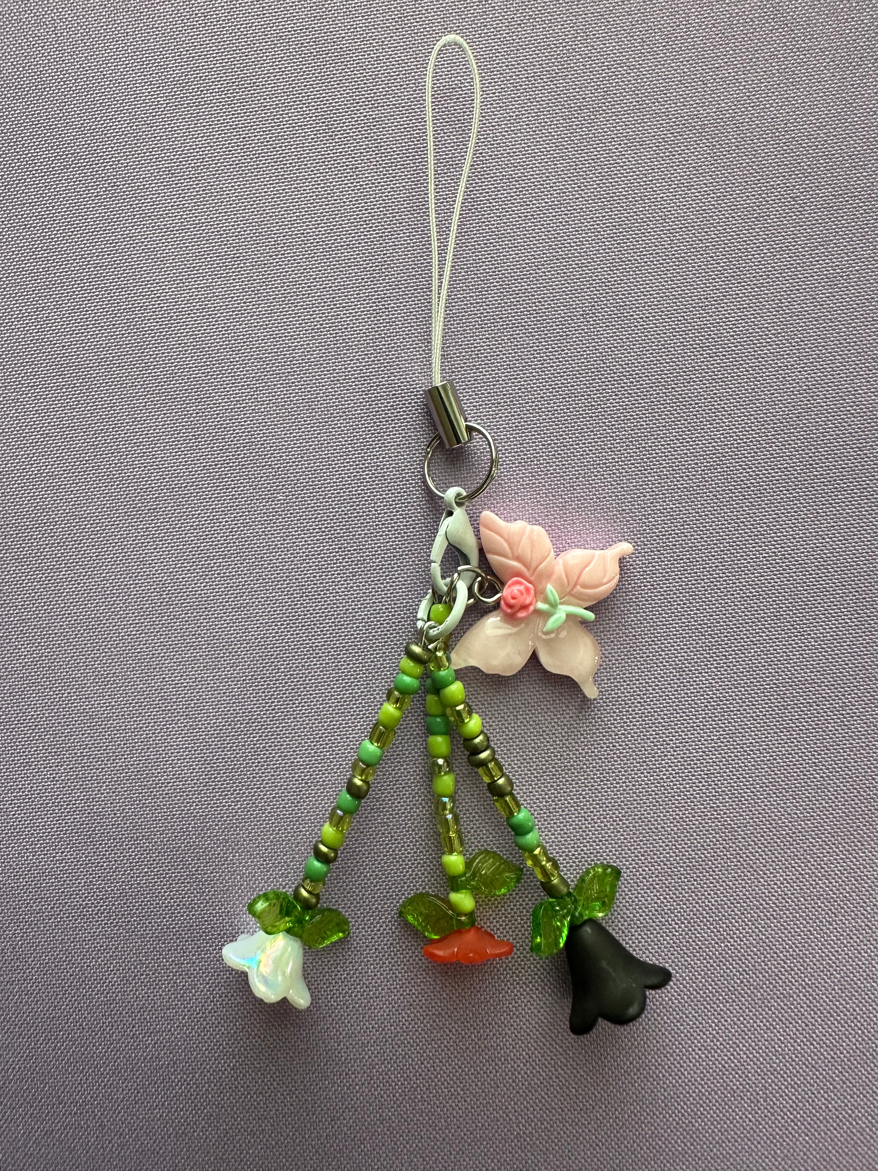 A handmade charm with a white loop strap, a silver clasp, a pink flower, and three dangling beaded stems with small flower charms in white, orange, and black, displayed on a textured gray background.