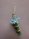 A decorative charm featuring a light blue translucent lotus flower and a green beaded handle with gold-tone metal accents, photographed on a textured mauve background.