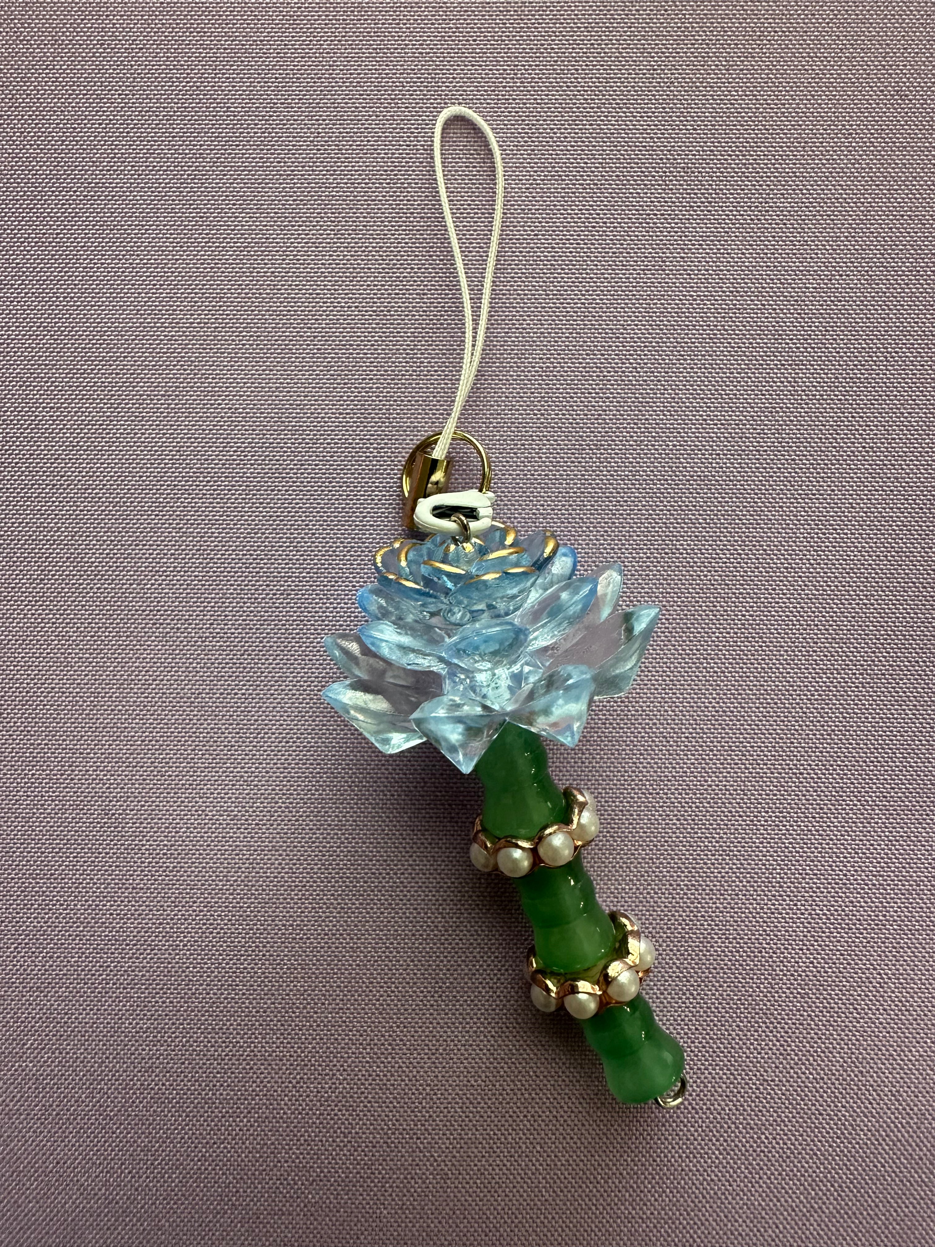 A decorative charm featuring a light blue translucent lotus flower and a green beaded handle with gold-tone metal accents, photographed on a textured mauve background.