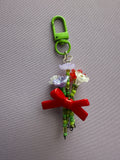 A handmade beaded keychain charm featuring a bright green plastic clasp, green beads, small white and lavender flowers, and a prominent red ribbon tied in a bow, displayed on a textured gray background.