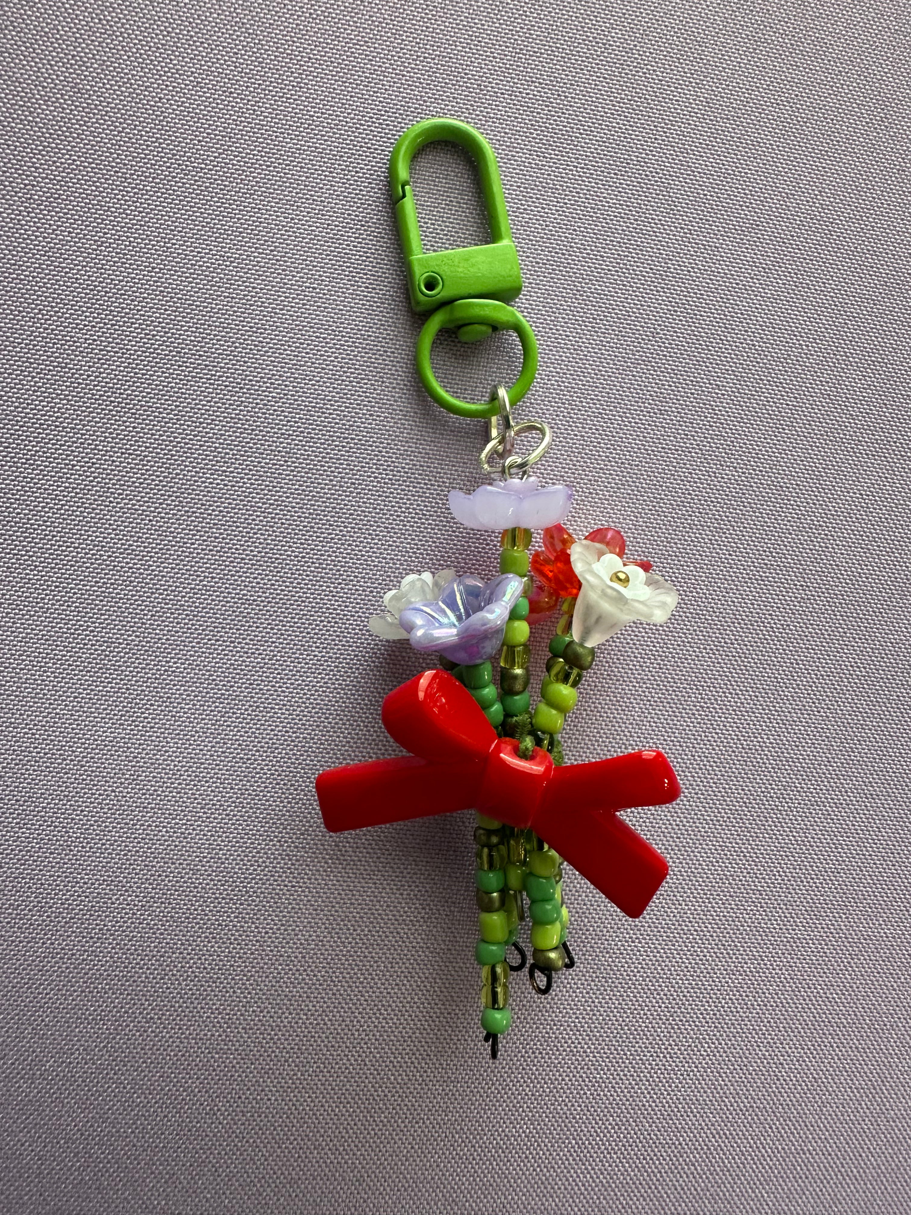 A handmade beaded keychain charm featuring a bright green plastic clasp, green beads, small white and lavender flowers, and a prominent red ribbon tied in a bow, displayed on a textured gray background.