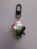 A decorative black carabiner keychain featuring a small floral arrangement with pink, white, and red flowers, green stems, and a black bow, displayed on a textured gray background.