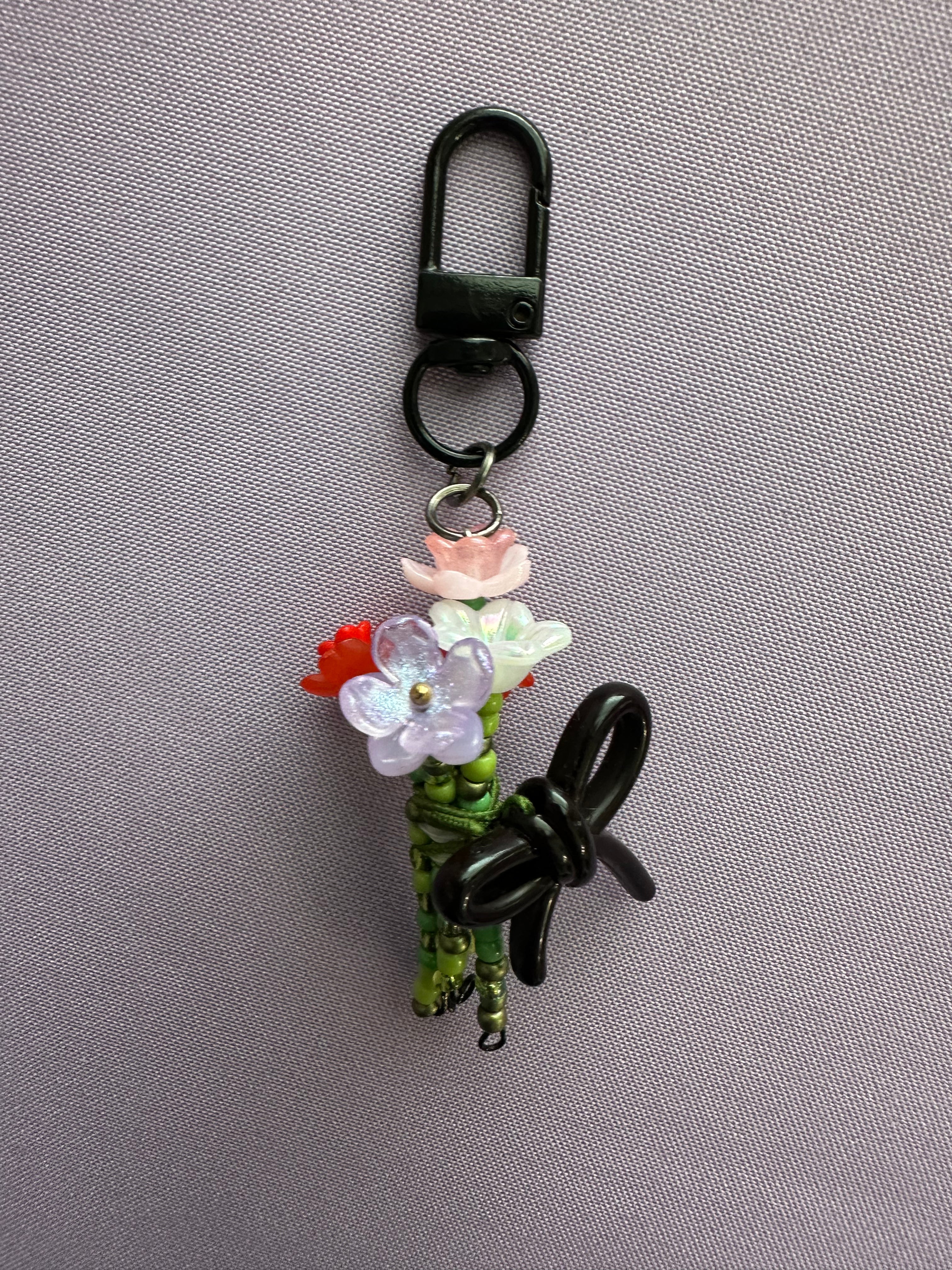 A decorative black carabiner keychain featuring a small floral arrangement with pink, white, and red flowers, green stems, and a black bow, displayed on a textured gray background.