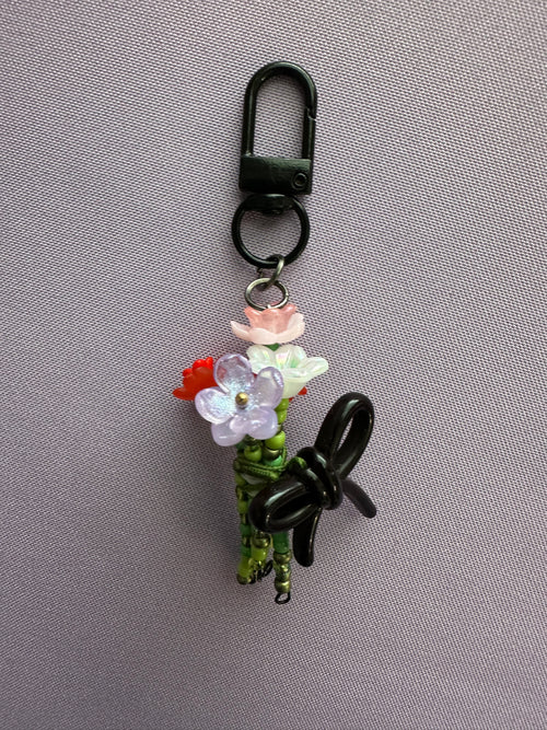 A decorative black carabiner keychain featuring a small floral arrangement with pink, white, and red flowers, green stems, and a black bow, displayed on a textured gray background.