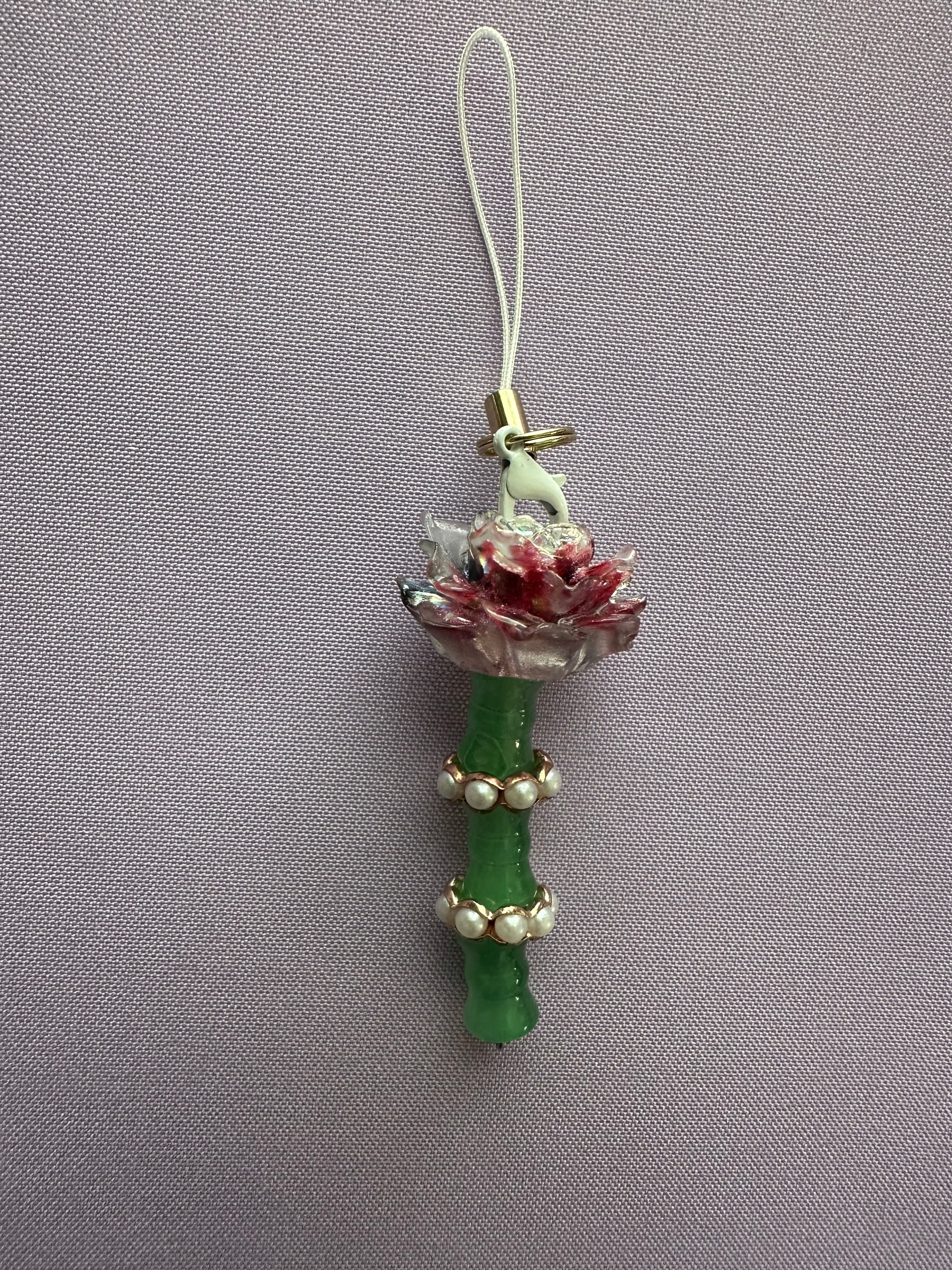 A decorative charm featuring a green bamboo stem with white pearl-like beads, a pink and white flower, a small white bird, and a white loop at the top, shown against a textured gray background.