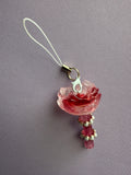 A pink rose charm with a white strap and gold-toned metal clasp, resting on a textured lavender-gray fabric background.