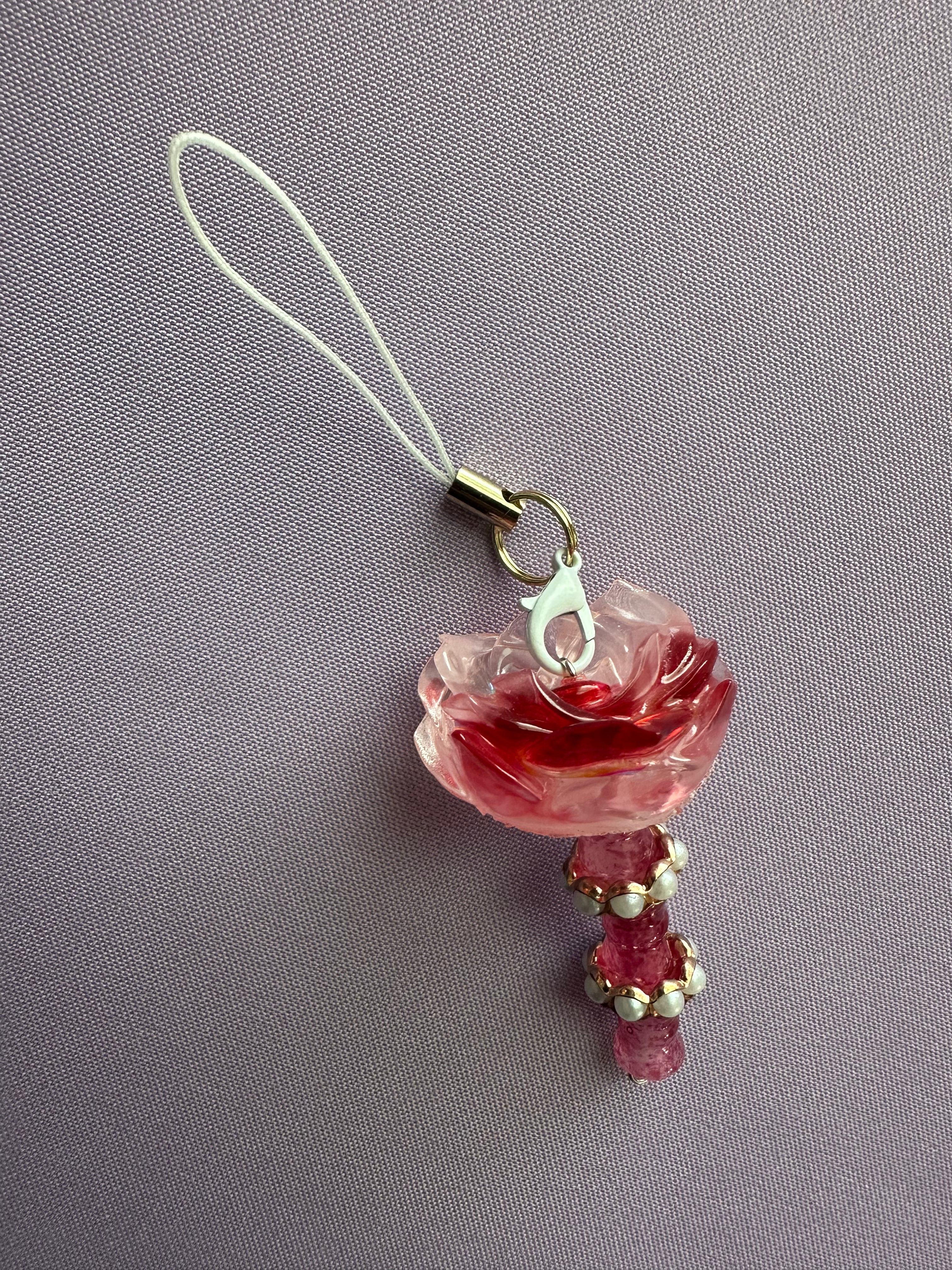 A pink rose charm with a white strap and gold-toned metal clasp, resting on a textured lavender-gray fabric background.