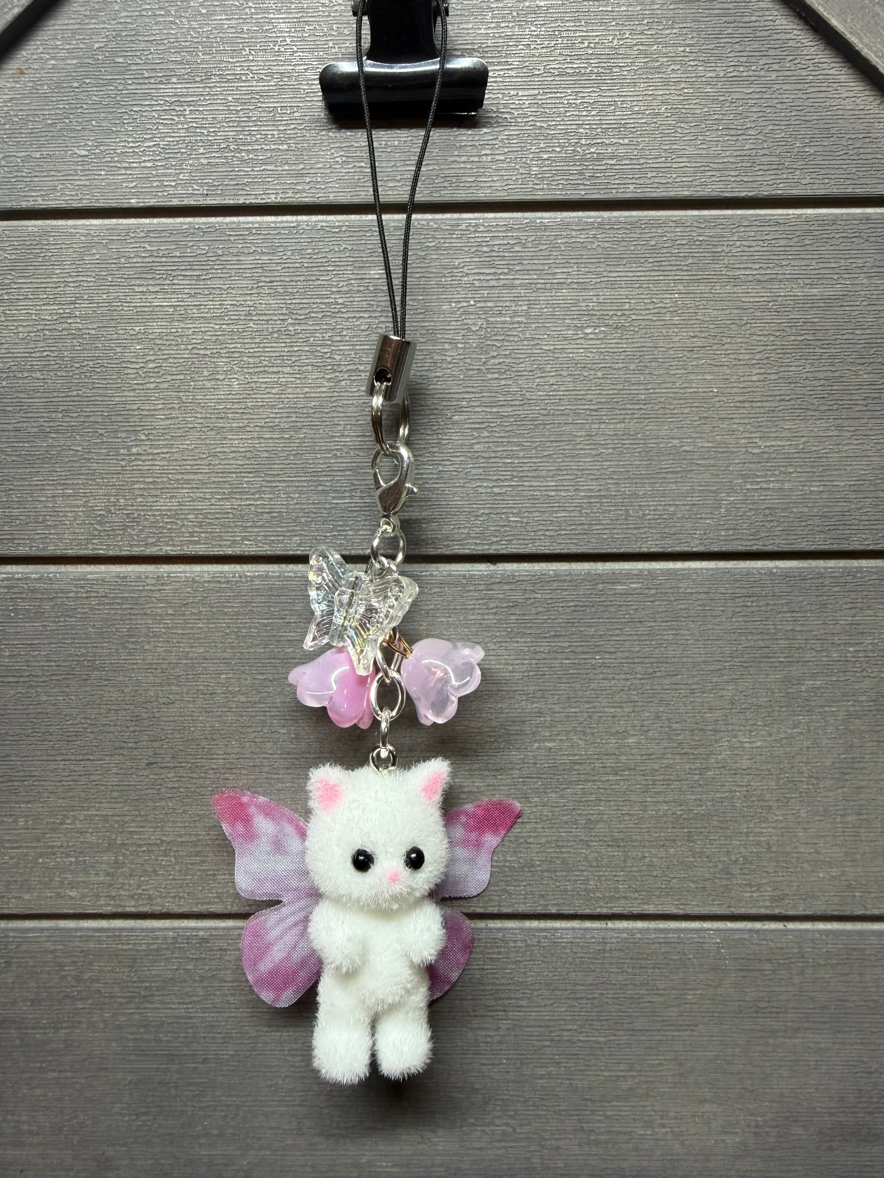 A white plush kitten charm with pink wings and small pink flower accents, hanging from a black cord on a gray wood plank background.
