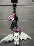 A gothic-themed charm featuring a white plush cat with bat wings, a pink cat charm, a tombstone charm, and a small bat charm, all attached to a black strap against a gray wood background.