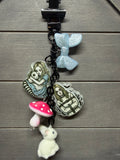 A black bag charm featuring Alice in Wonderland themed charms, including a light blue glittery bow, a pink and white mushroom, a small white bunny, and two illustrated character pendants, displayed on a gray wood background.