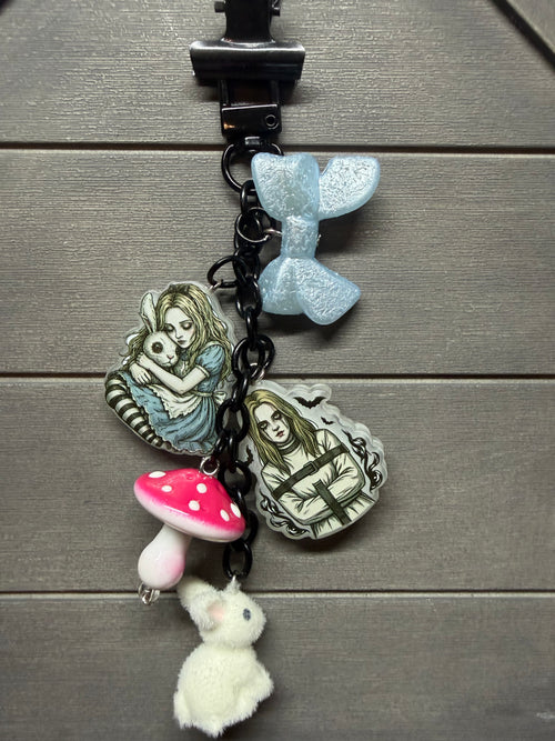 A black bag charm featuring Alice in Wonderland themed charms, including a light blue glittery bow, a pink and white mushroom, a small white bunny, and two illustrated character pendants, displayed on a gray wood background.
