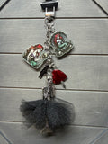 A gothic-themed keychain featuring a skeleton figure in a black tulle tutu, a red heart-shaped charm, and two fairy charms with green and red enamel details, all attached to a silver chain.