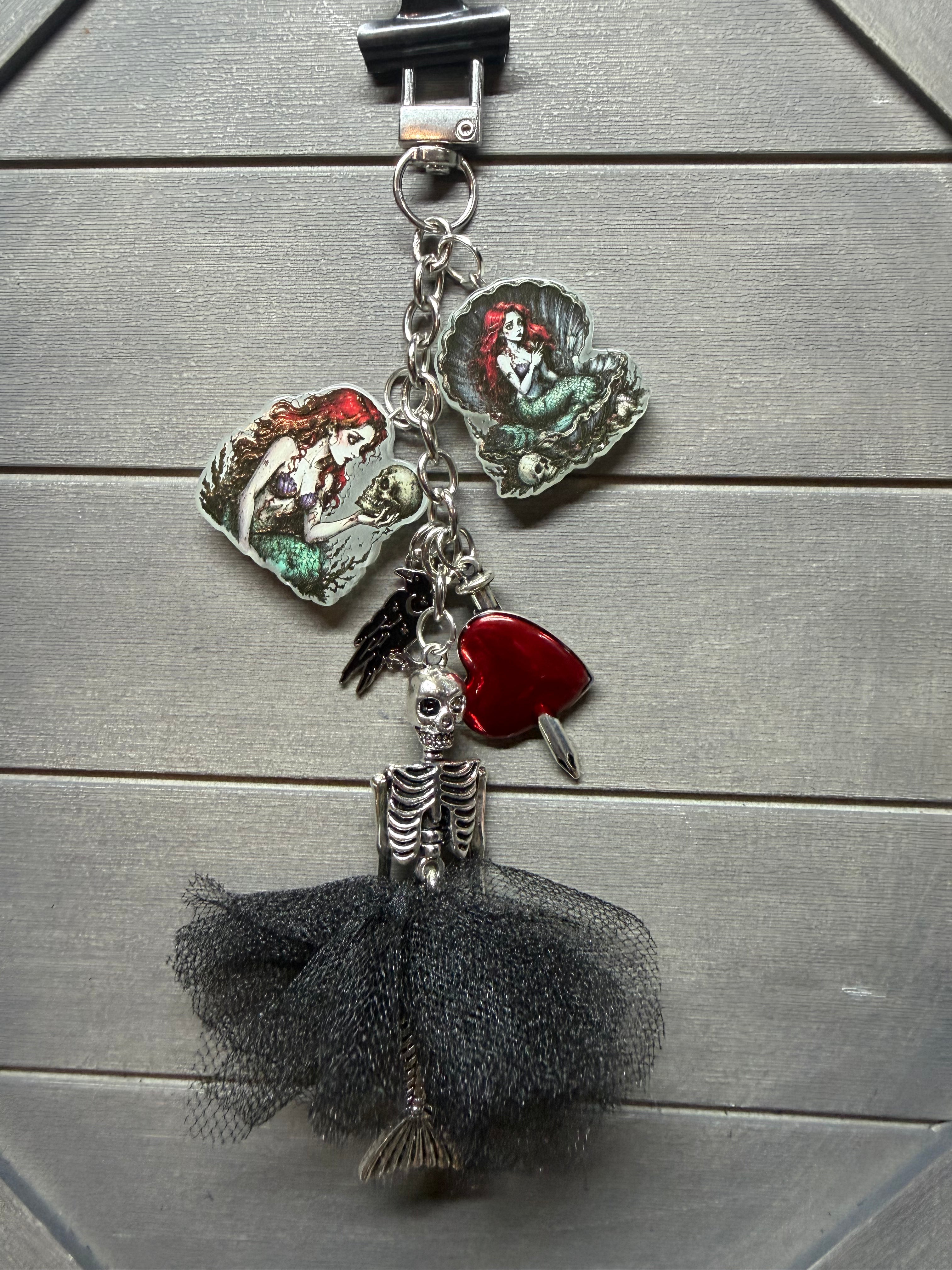 A gothic-themed keychain featuring a skeleton figure in a black tulle tutu, a red heart-shaped charm, and two fairy charms with green and red enamel details, all attached to a silver chain.
