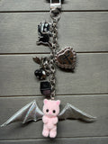 A gothic-style silver chain keychain featuring a pink plush bat character with silver wings, a silver heart charm with an anchor, a black cat charm, and a small black bead bell, displayed on a gray wood plank background.