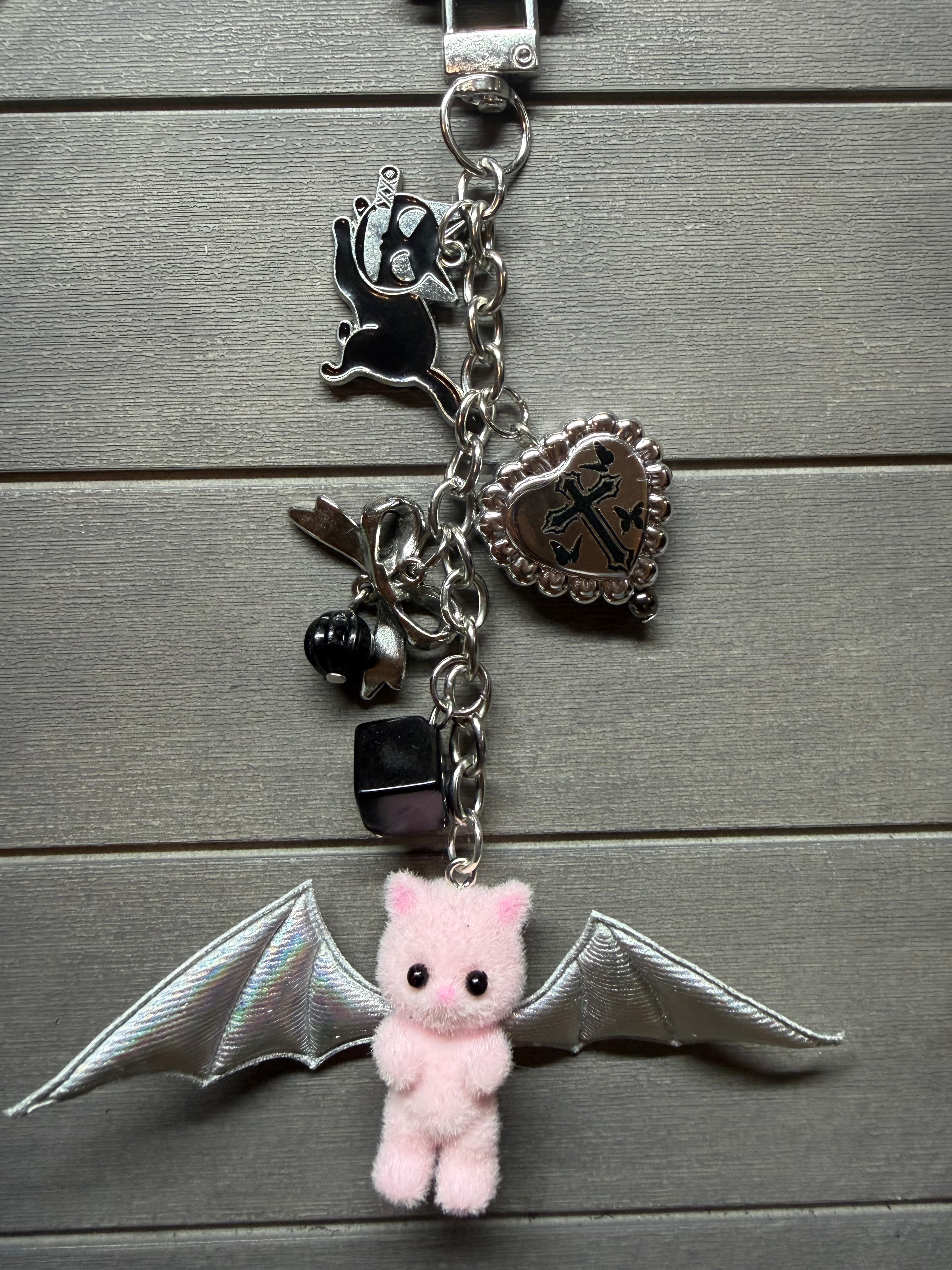 A gothic-style silver chain keychain featuring a pink plush bat character with silver wings, a silver heart charm with an anchor, a black cat charm, and a small black bead bell, displayed on a gray wood plank background.