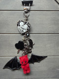A gothic-themed keychain featuring a red felt bat-shaped teddy bear, a silver heart charm with a skeleton and rhinestones, a black heart charm, and a black bat wing charm, displayed on a gray wood background.