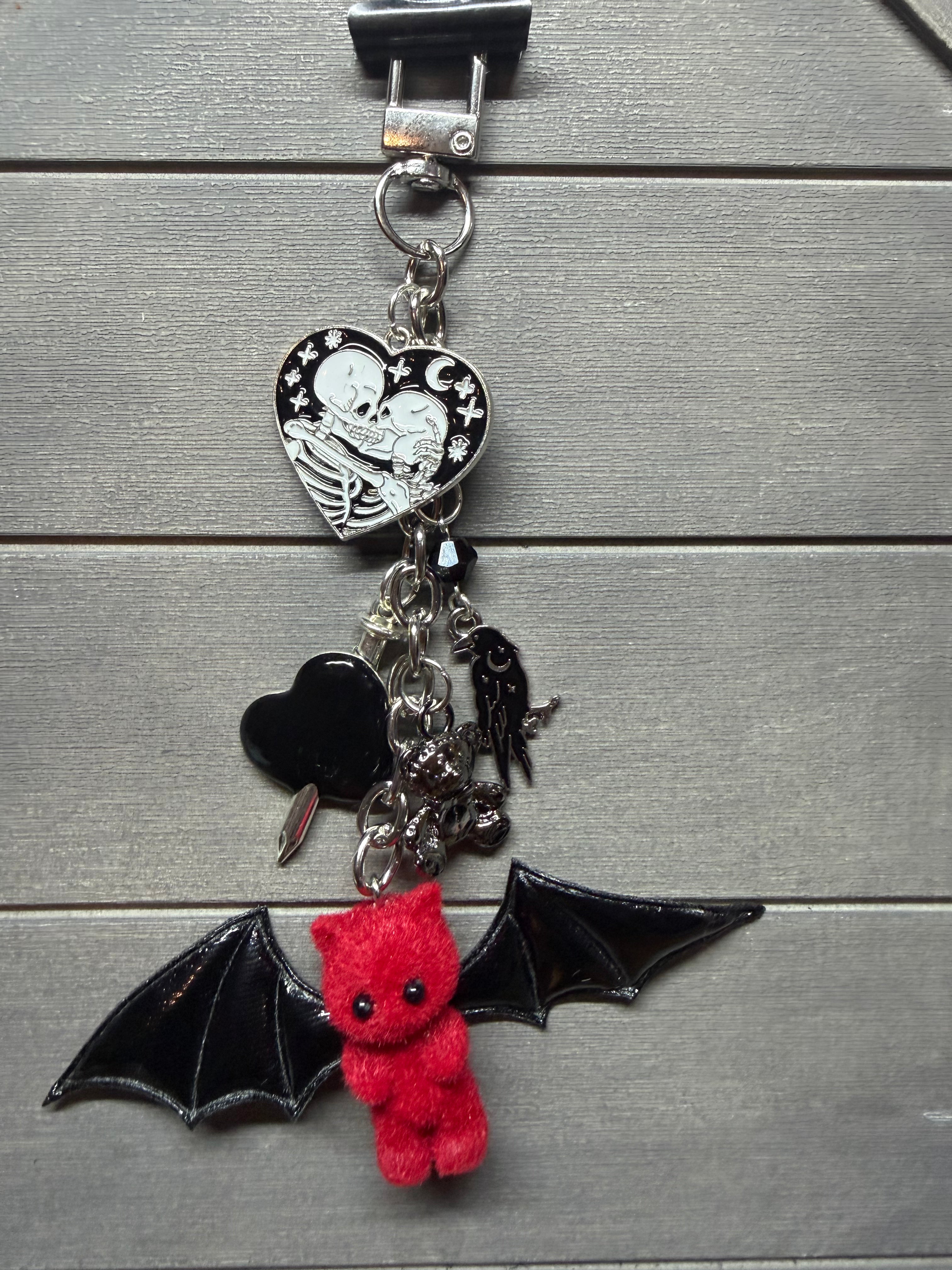 A gothic-themed keychain featuring a red felt bat-shaped teddy bear, a silver heart charm with a skeleton and rhinestones, a black heart charm, and a black bat wing charm, displayed on a gray wood background.