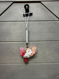 A handmade floral charm with pink, white, and peach flowers, a red leaf, and a braided white cord, hanging from a black clip against a gray wood-paneled background.