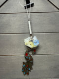 A decorative peacock phone or keychain charm with a white braided cord, small white and light blue flowers, and a dark metal peacock body accented with red and turquoise details, shown against a gray wood plank background.