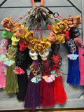 A set of colorful Día de los Muertos skull charms hanging from a gray wooden surface. Each keychain features a skull charm, dried roses, a tassel, and various charms including crystals and chains.