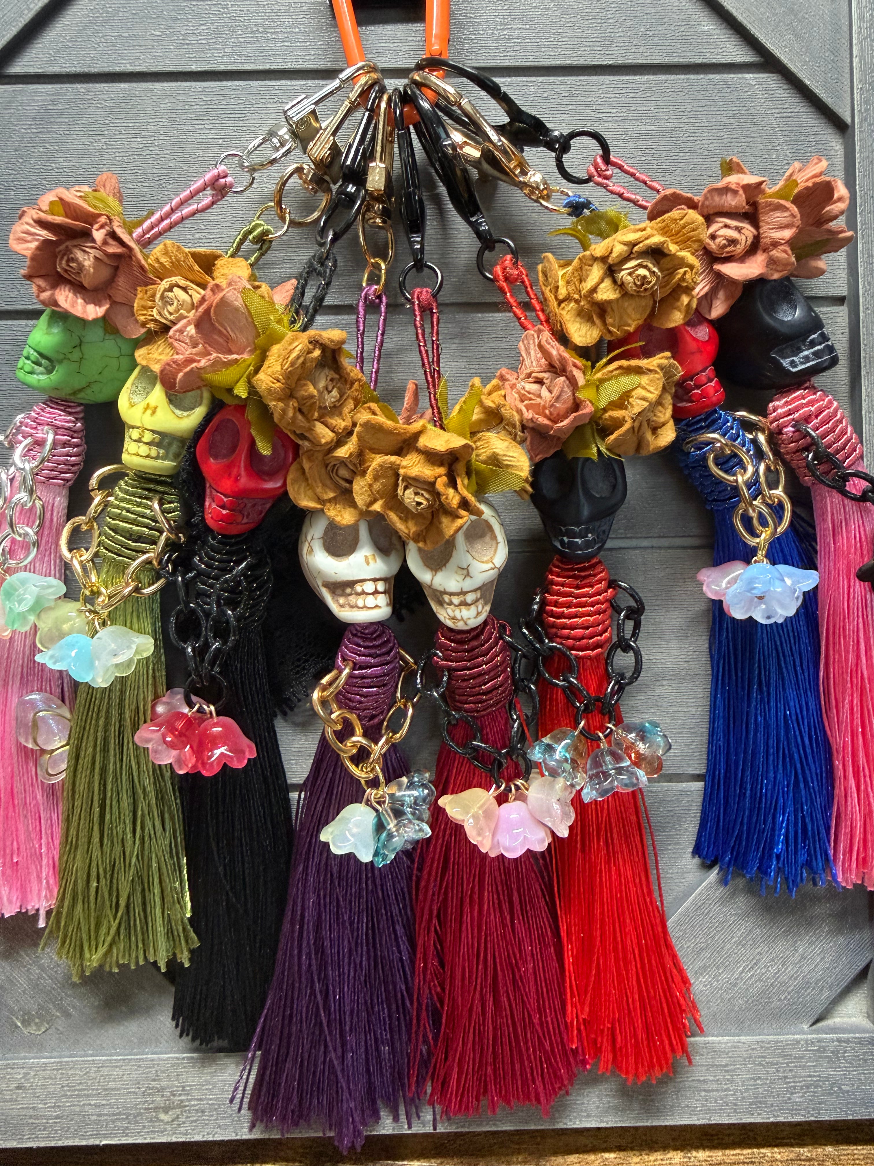 A set of colorful Día de los Muertos skull charms hanging from a gray wooden surface. Each keychain features a skull charm, dried roses, a tassel, and various charms including crystals and chains.