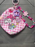 A pink and white gingham Sanrio My Melody keychain pouch with a matching pink beaded wrist strap, resting on a dark gray wooden surface.