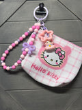 A pink and white Hello Kitty zippered coin purse with a matching beaded wrist strap and character charms, displayed on a dark gray wood background.