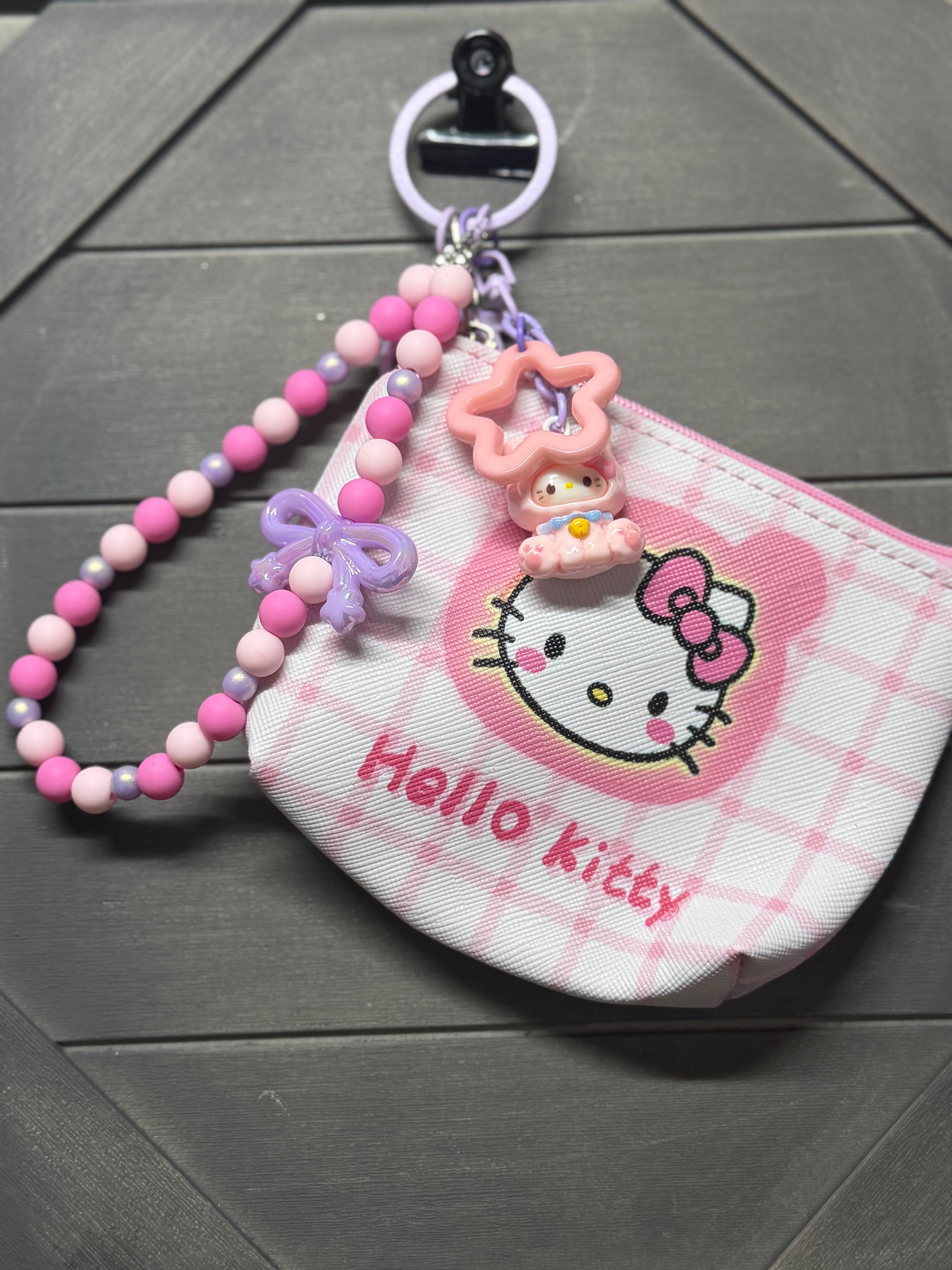 A pink and white Hello Kitty zippered coin purse with a matching beaded wrist strap and character charms, displayed on a dark gray wood background.