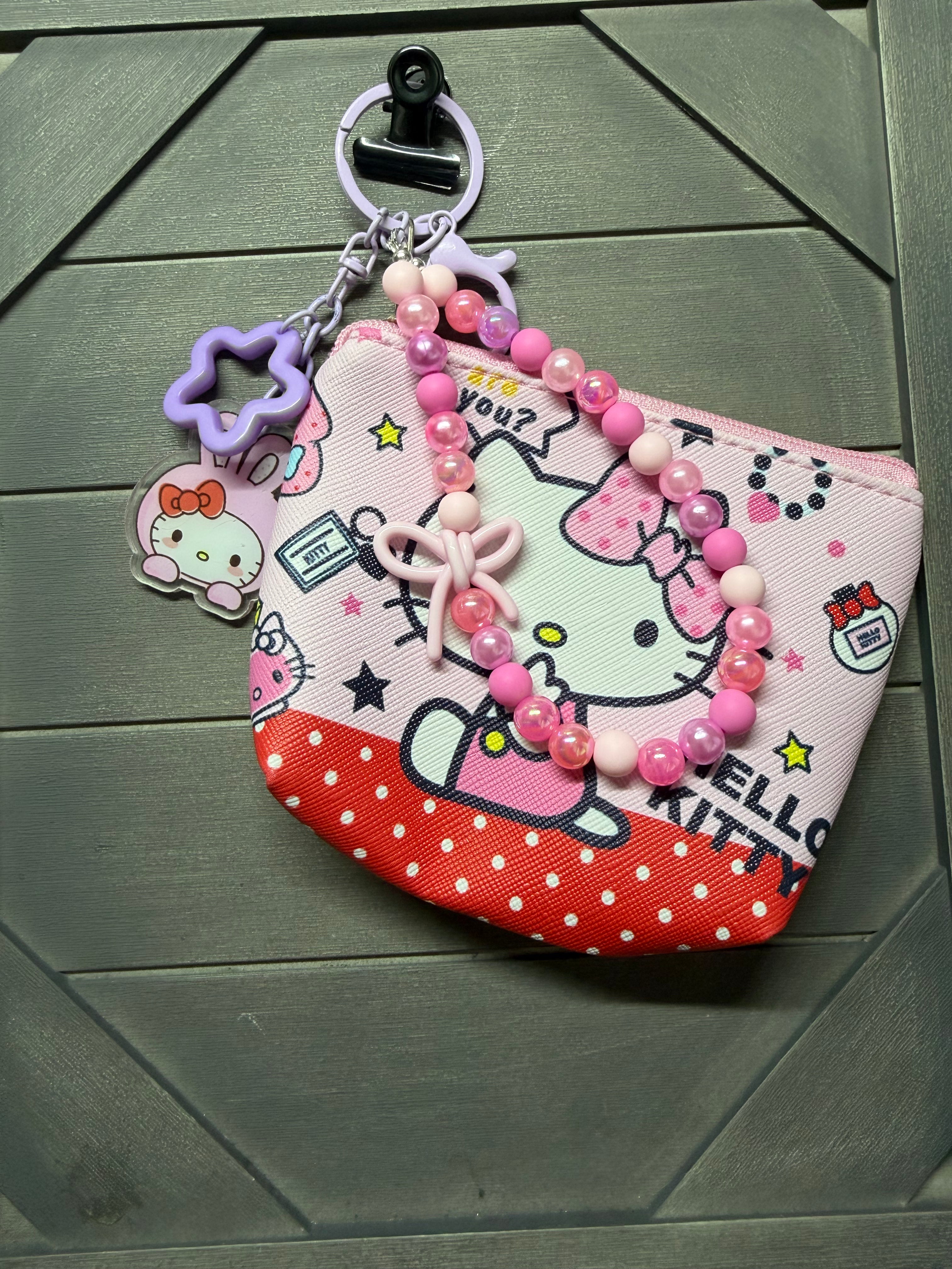 A pink and red Hello Kitty zippered pouch with a pink beaded bracelet and a purple flower charm, displayed on a dark green wood background.