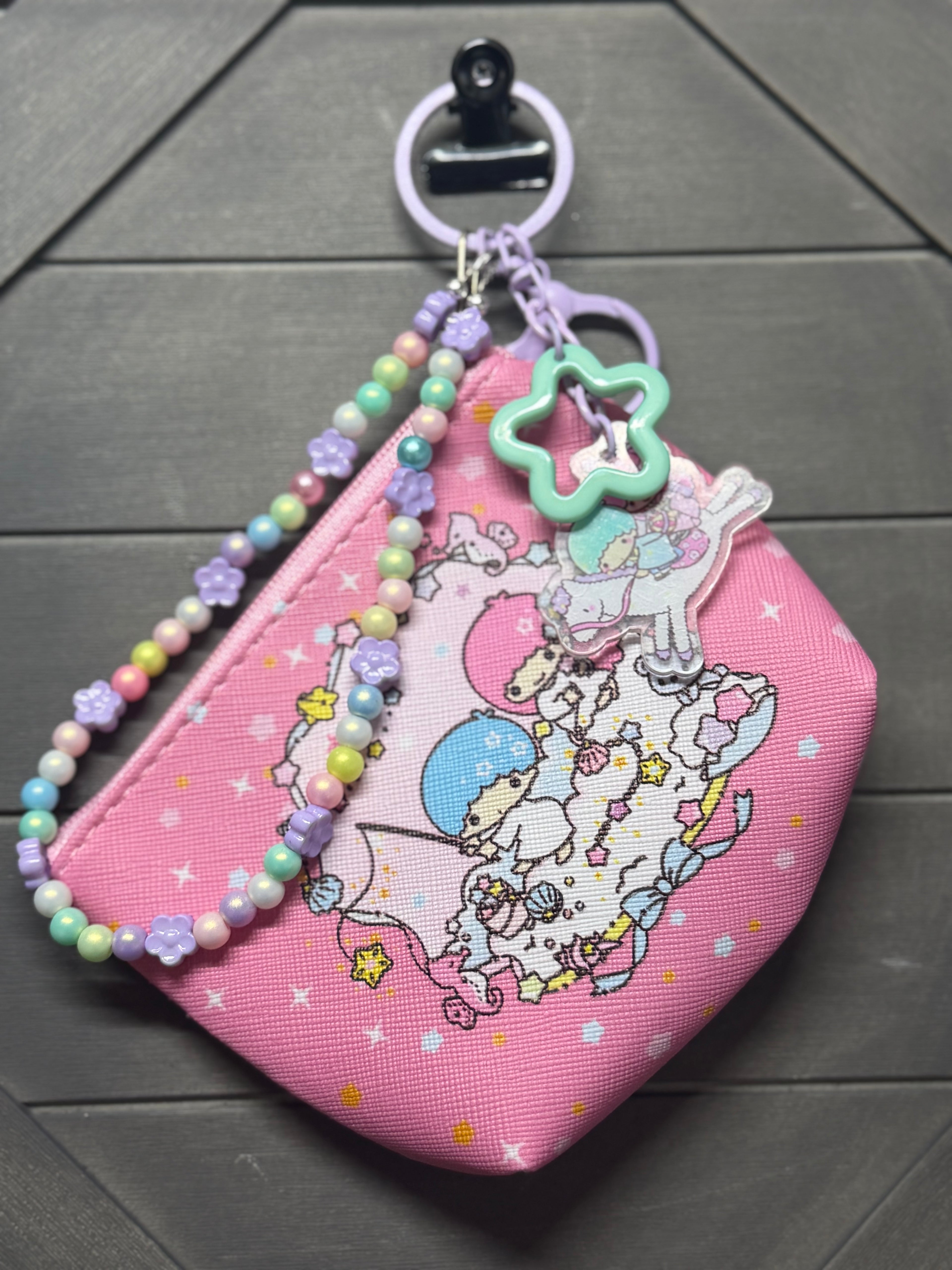 A pink fabric coin purse with a cartoon sea print, decorated with pastel beaded wrist straps, a purple key ring, and a green star charm, shown on a dark gray wood background.