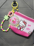 A pink and white Hello Kitty zippered coin purse with a colorful beaded wrist strap, a flower charm, and a round character medallion, displayed on a dark gray wood-textured background.