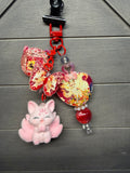 A colorful purse charm featuring red and gold dragon motifs, a pink fluffy fox character, and red beads, displayed on a gray wood background.