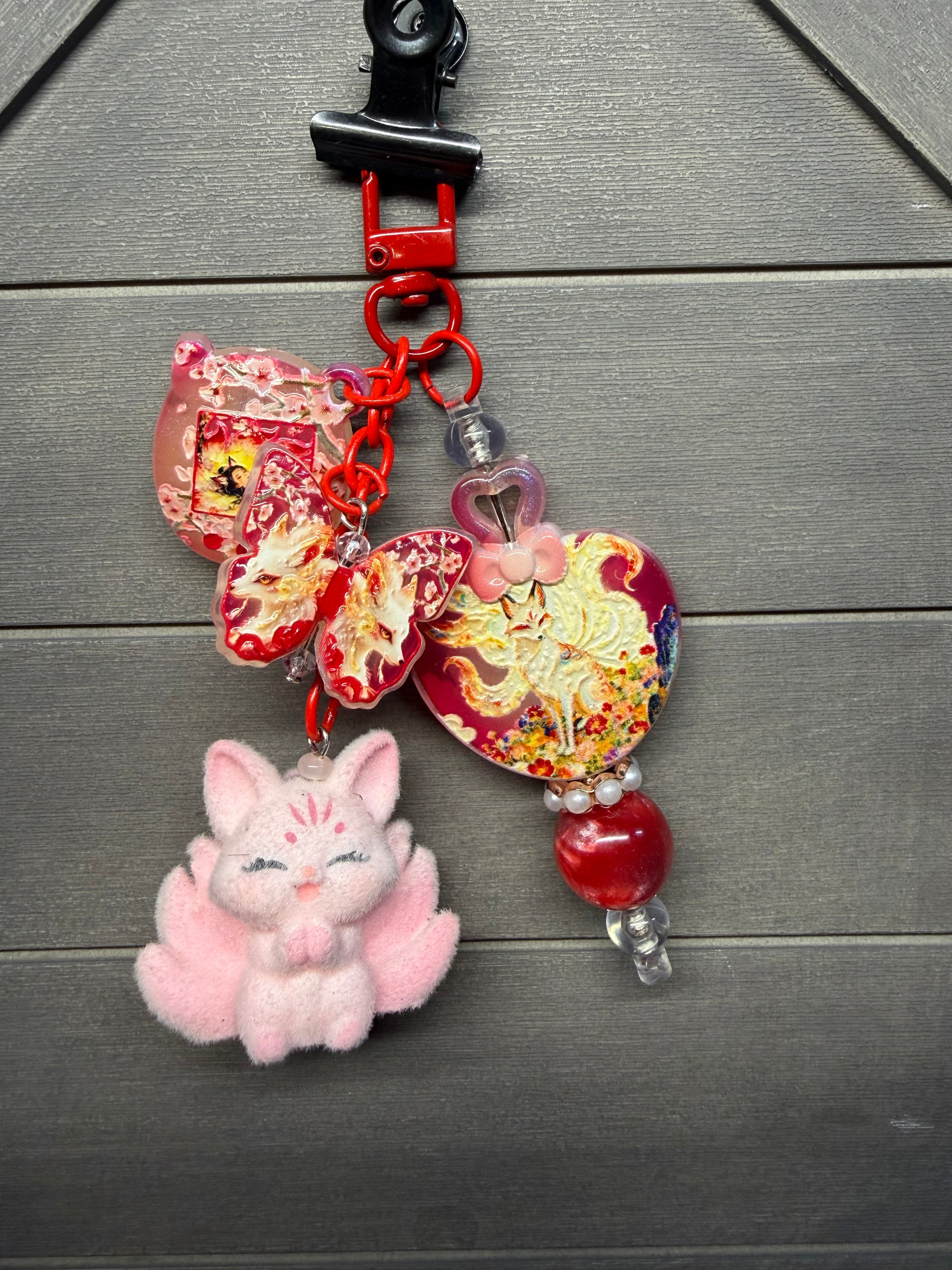 A colorful purse charm featuring red and gold dragon motifs, a pink fluffy fox character, and red beads, displayed on a gray wood background.