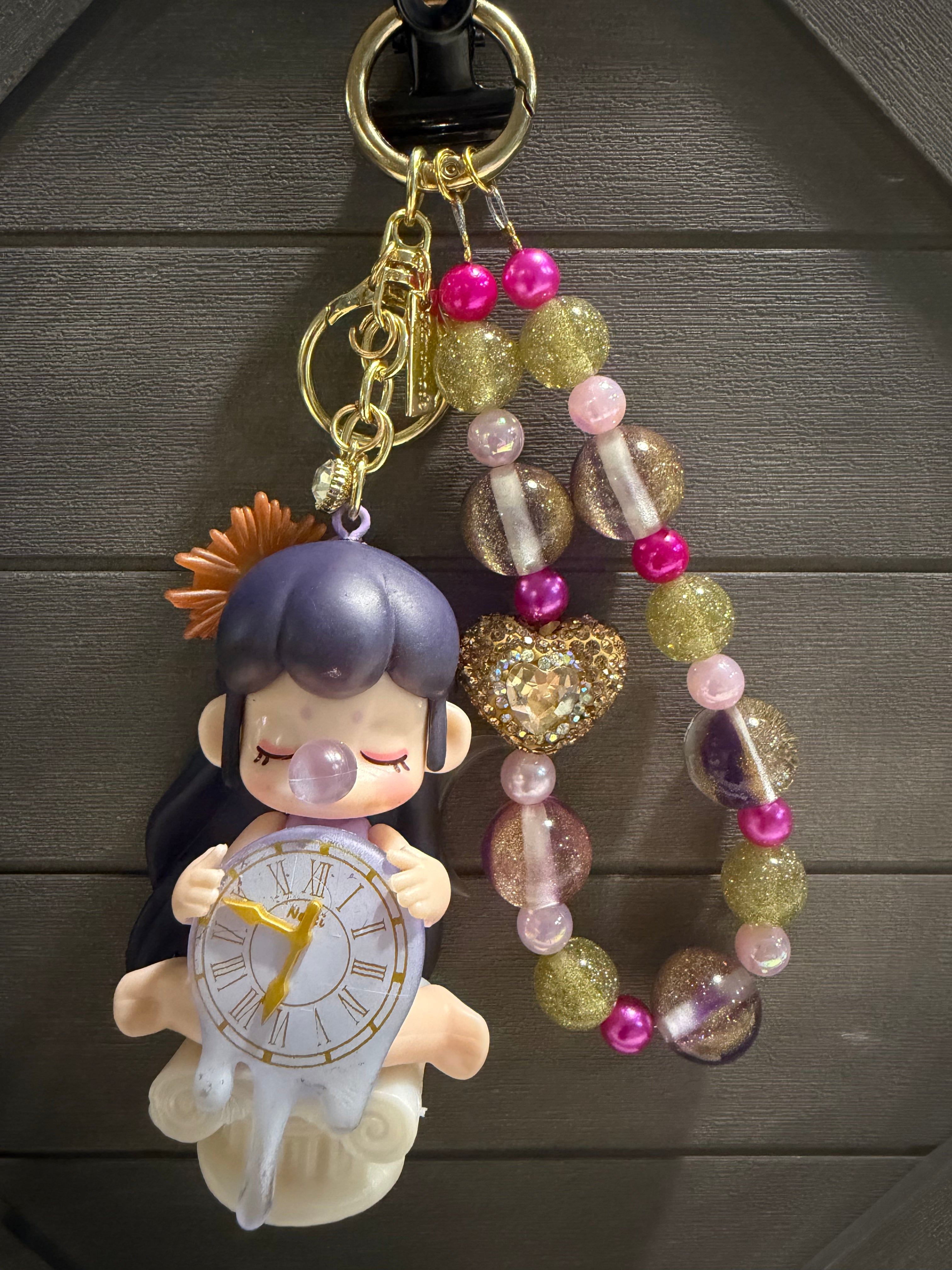 A decorative charm hanging against a dark wood background. The set includes a pastel anime-style character holding a clock, gold-tone rings and chain, translucent and glittery beads in pink, lavender, and gold, and a small orange flower accent.