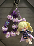 A handmade beaded charm featuring purple and lavender roses, purple beads, a lavender bow, and a cute blonde anime doll charm holding a small pink bunny, hanging from a silver heart-shaped clasp against a dark gray wooden background.