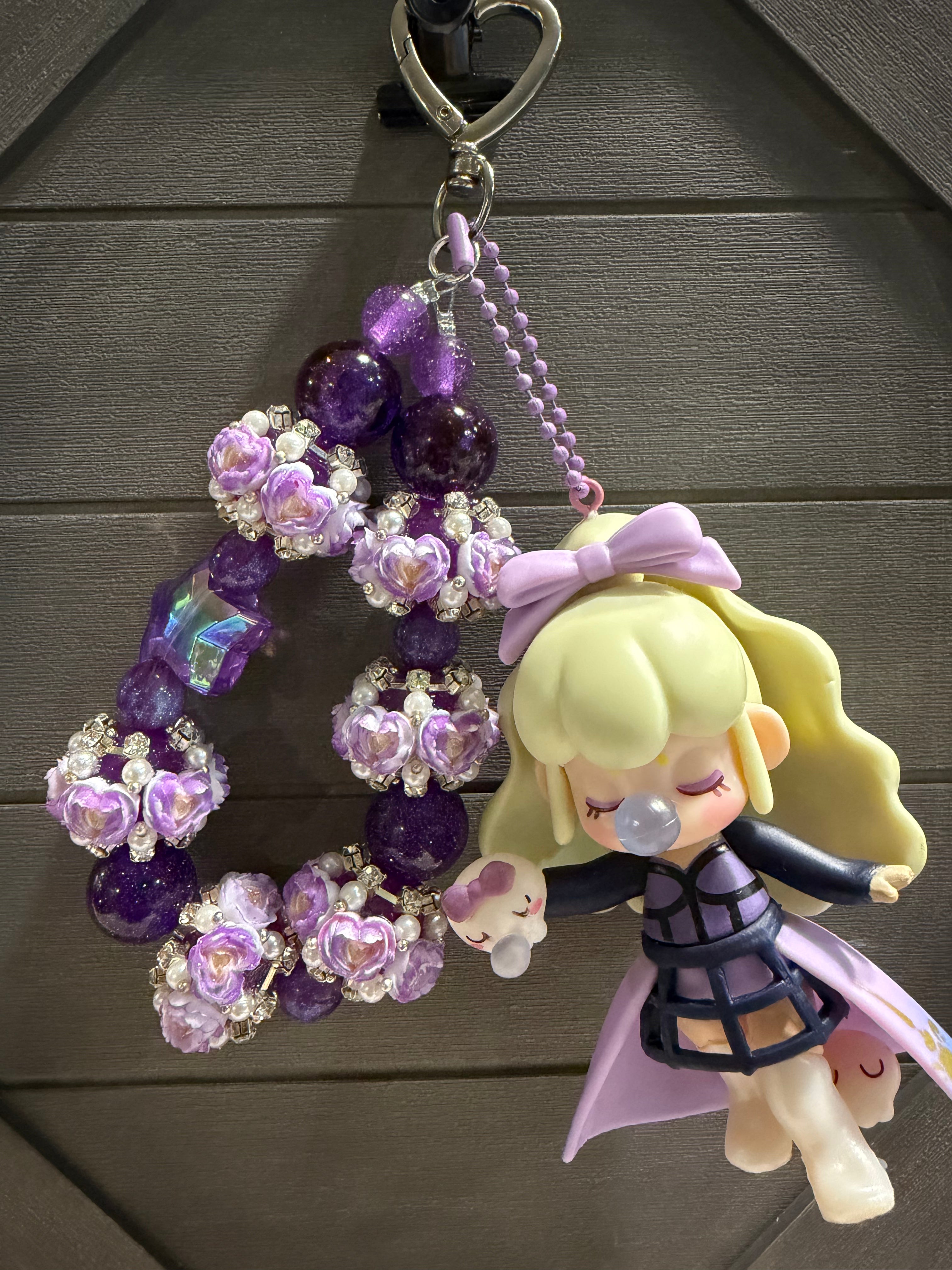 A handmade beaded charm featuring purple and lavender roses, purple beads, a lavender bow, and a cute blonde anime doll charm holding a small pink bunny, hanging from a silver heart-shaped clasp against a dark gray wooden background.