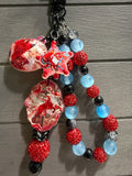 A beaded charm featuring red glittery beads, light blue beads, black beads, and two large marbled red and white glass charms on a gray wood background.