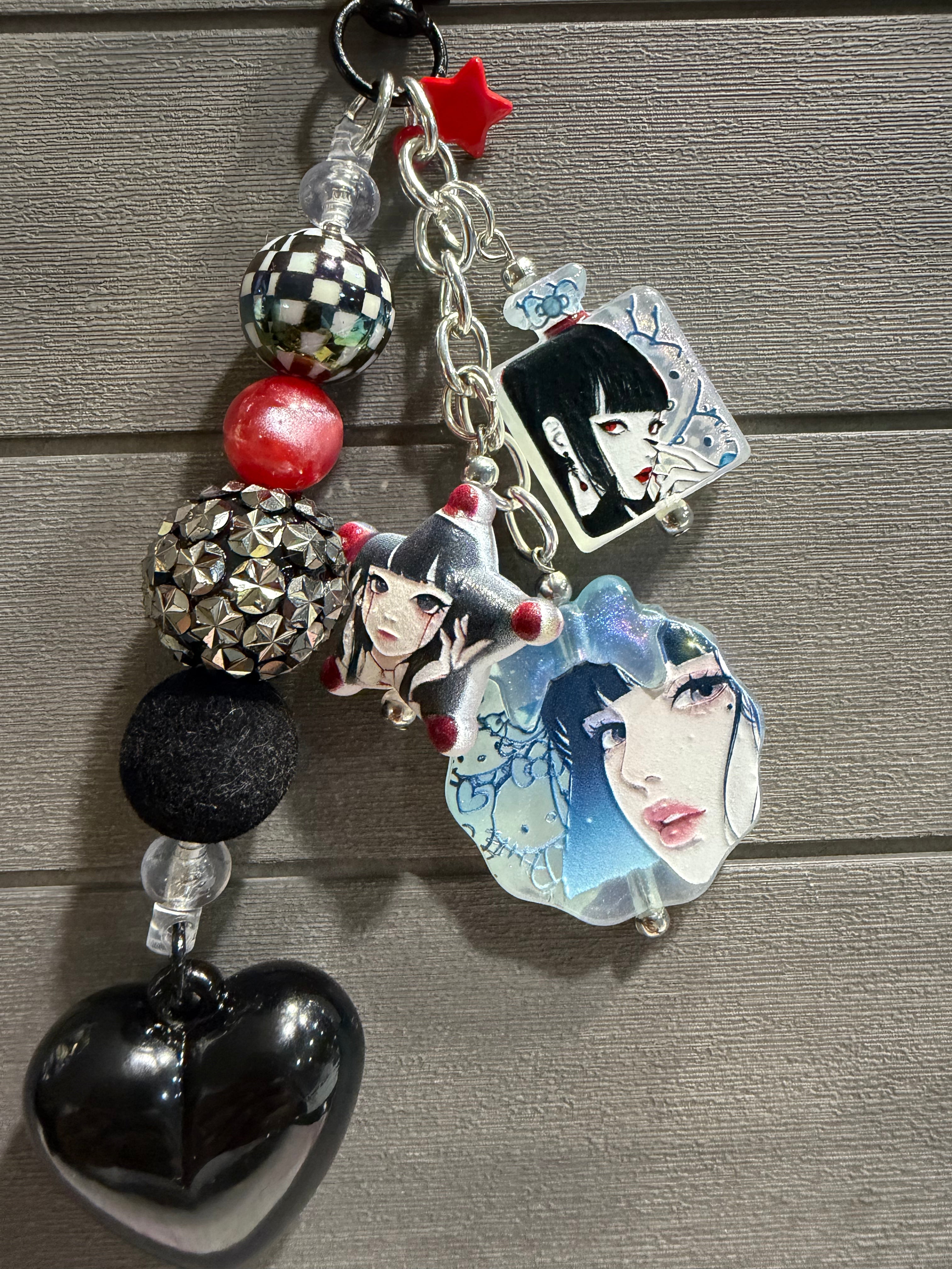 A gothic-themed charm featuring anime-style character charms, a black heart-shaped bead, patterned and metallic beads, and a silver chain, displayed on a gray wood background.