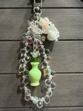 A decorative charm with a small white bunny figure, clear faceted beads, an iridescent butterfly charm, a light green vase, and colorful rhinestone accents, hanging against a dark gray wood plank background.