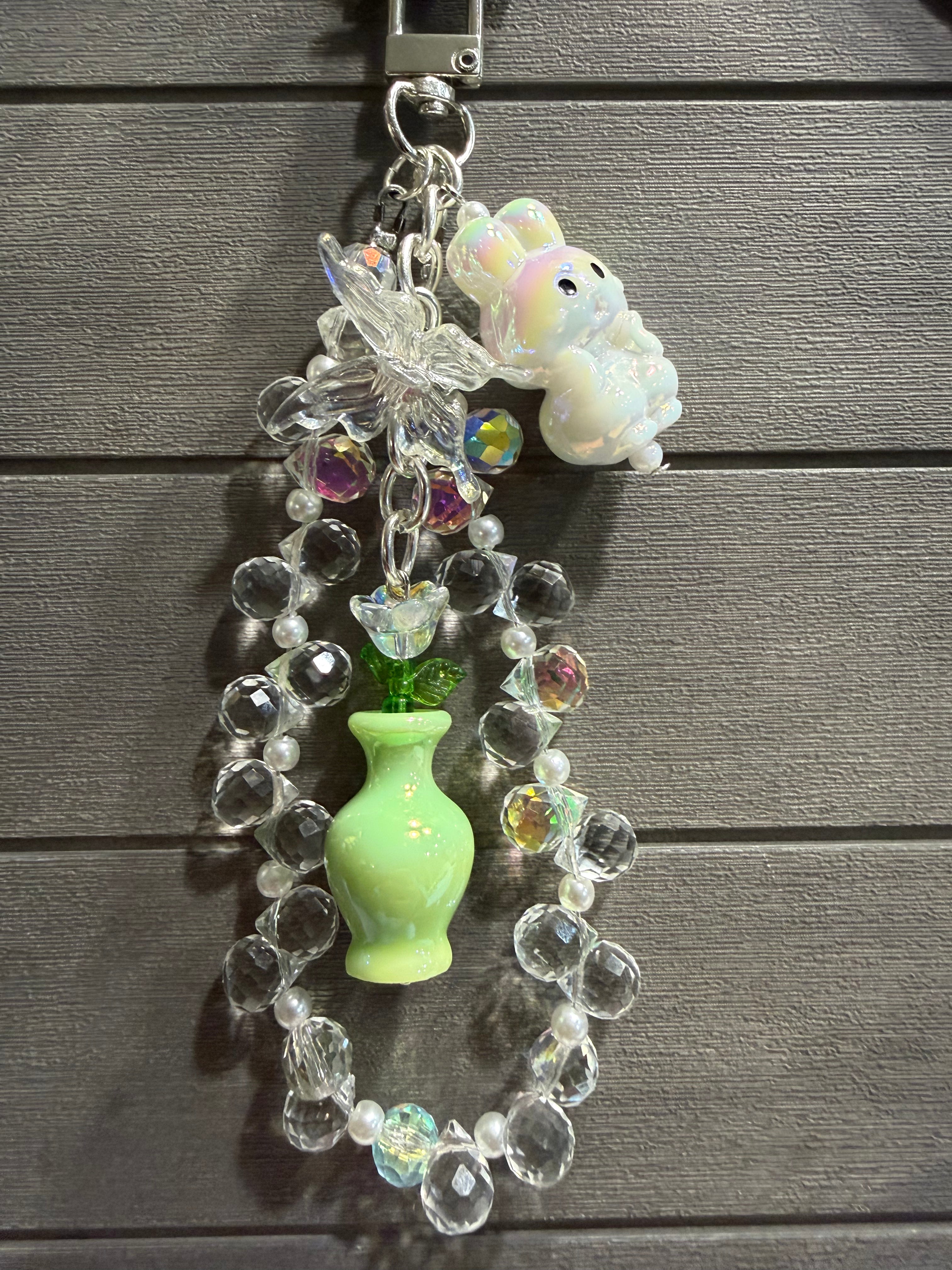 A decorative charm with a small white bunny figure, clear faceted beads, an iridescent butterfly charm, a light green vase, and colorful rhinestone accents, hanging against a dark gray wood plank background.