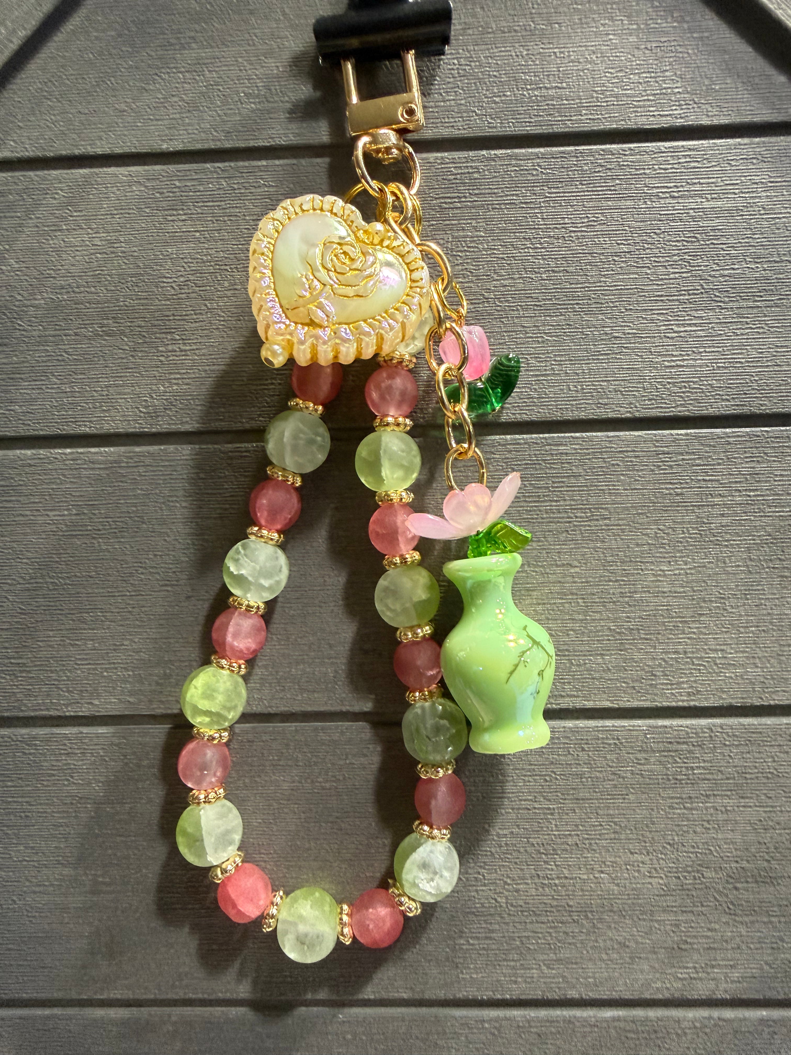 A decorative charm with a gold-tone clasp, a heart-shaped charm, faceted beads in pink and light green, a small pink flower, and a green vase charm, displayed against a dark gray wood-grain background.