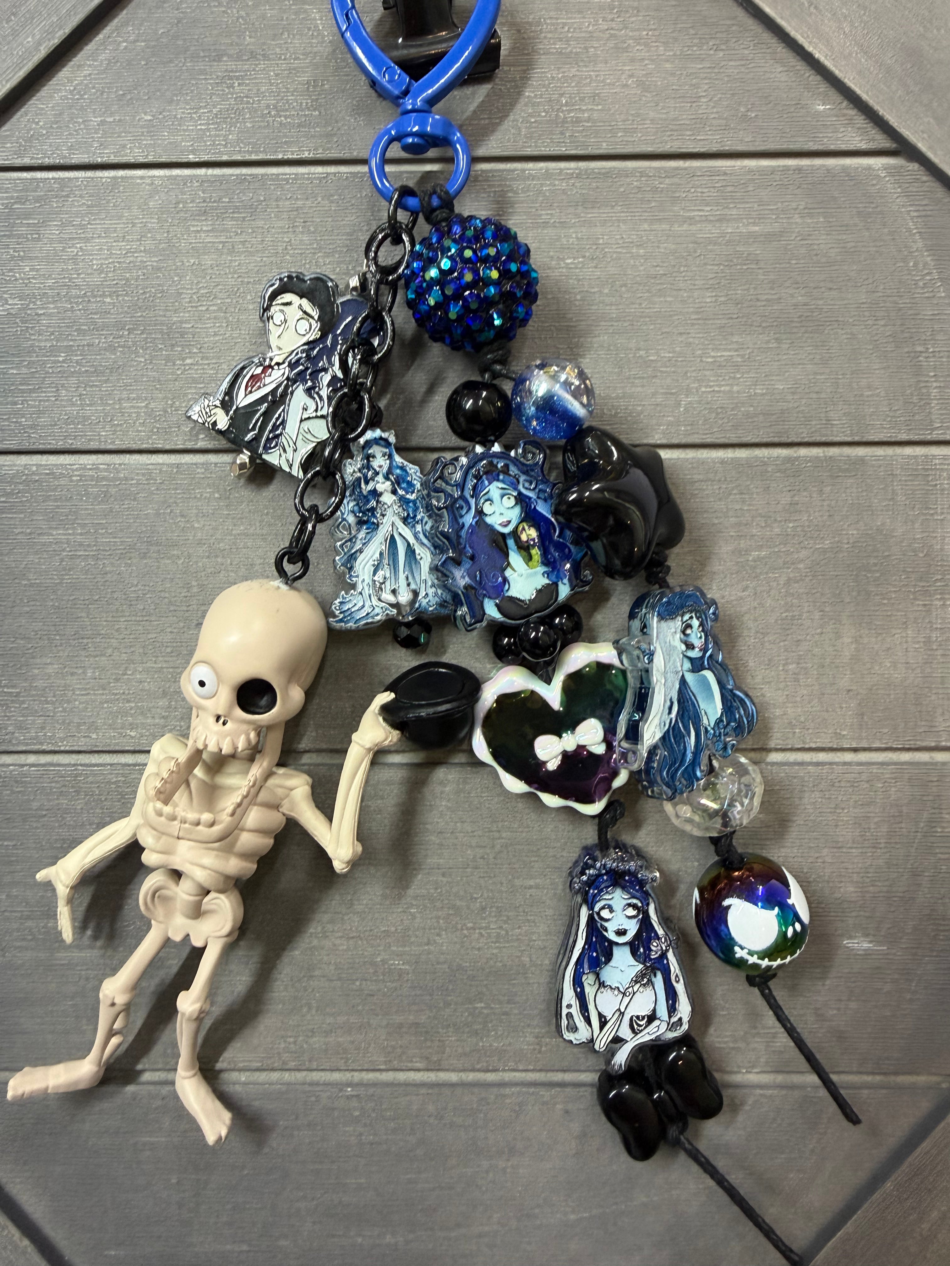 A gothic-themed keychain featuring a small skeleton figurine, blue and black beads, glass charms with illustrated female characters, and a blue metal key ring, displayed on a gray wood background.