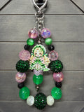 A beaded keychain featuring a central anime-style character charm surrounded by green, pink, black, and iridescent beads, displayed on a gray wood background.
