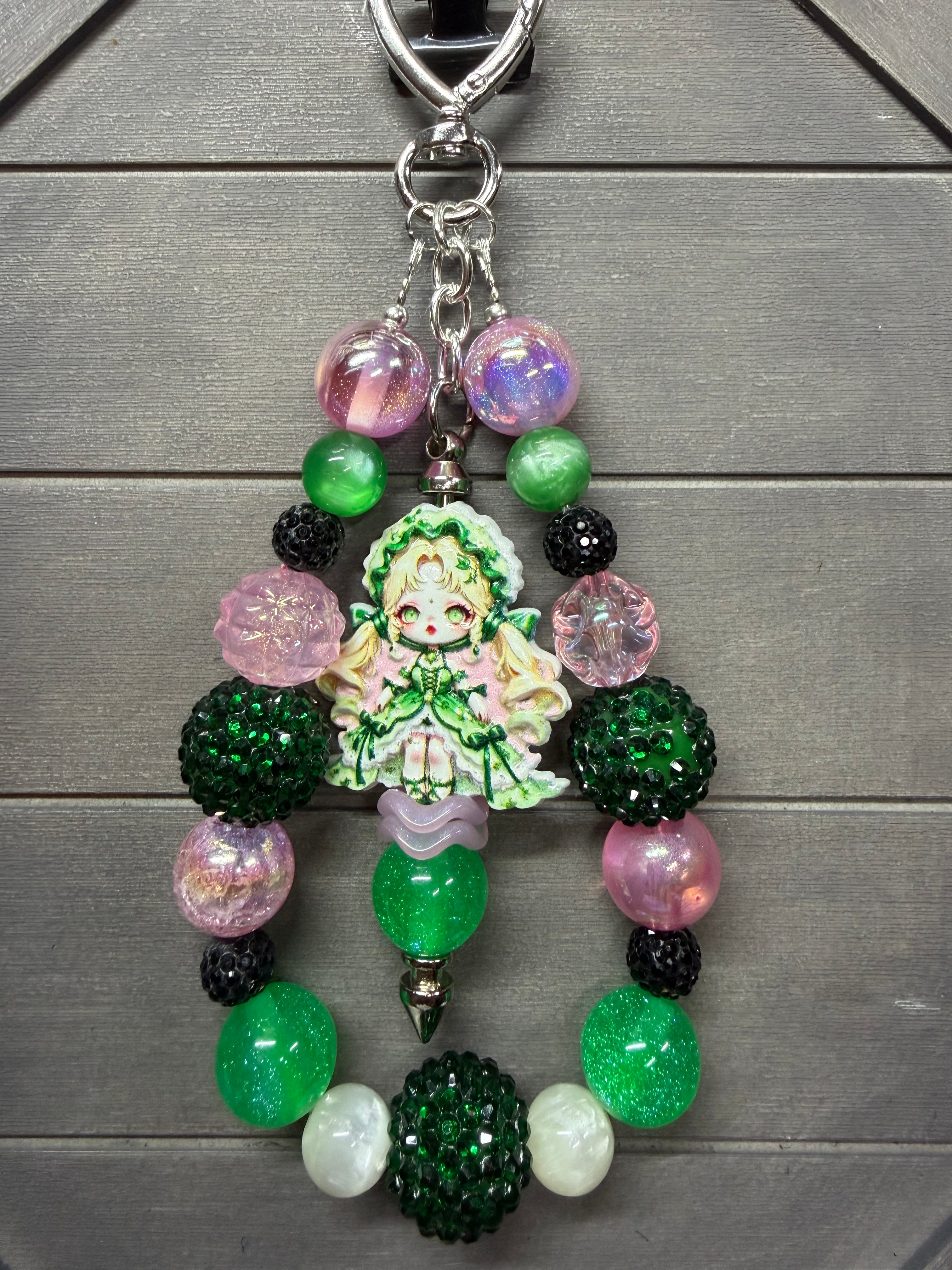 A beaded keychain featuring a central anime-style character charm surrounded by green, pink, black, and iridescent beads, displayed on a gray wood background.