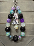 A beaded charm featuring a central white porcelain doll charm, black textured beads, iridescent purple and blue beads, and a silver metal ring, displayed on a gray wooden background.