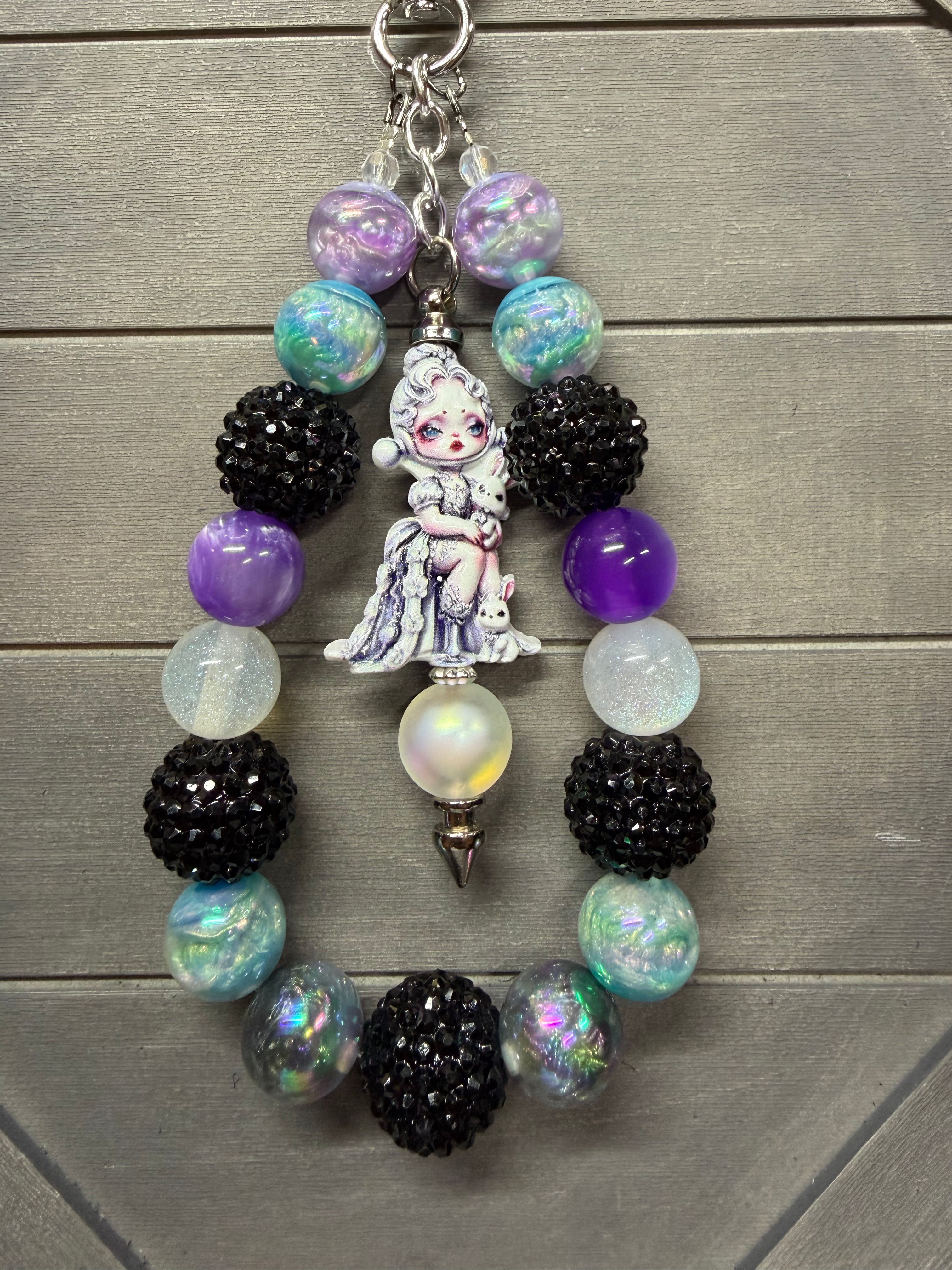 A beaded charm featuring a central white porcelain doll charm, black textured beads, iridescent purple and blue beads, and a silver metal ring, displayed on a gray wooden background.